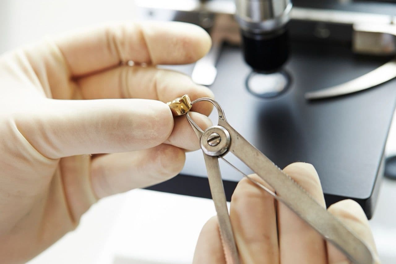A photo of gloved hands trimming a gold cap with an instrument.